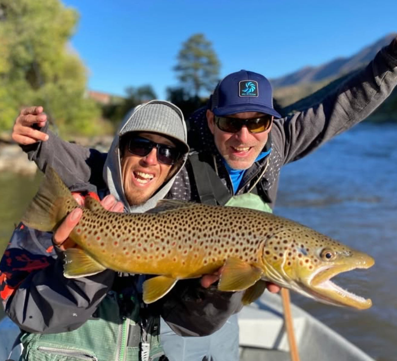 Two people holding a large brown trout by a body of water with trees and mountains in the background.