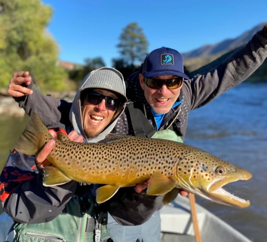 Two people holding a large brown trout by a body of water with trees and mountains in the background.