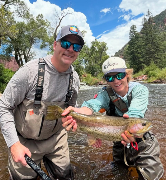 Two men holding a large fish by a river with trees and mountains in the background
