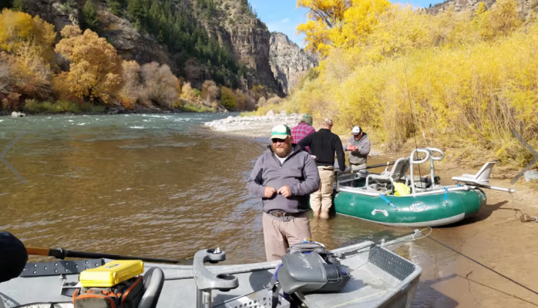 People fishing by a river with a green boat and mountains in the background